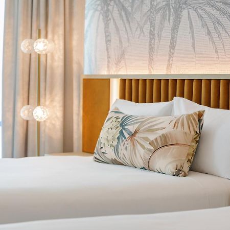 Amora Hotel Брисбен Экстерьер фото The photo shows a stylish hotel room featuring two neatly made beds with white linens. Each bed has several pillows, one of which has a decorative cover with a tropical plant pattern. Behind the beds is a modern headboard, and the wall is decorated w