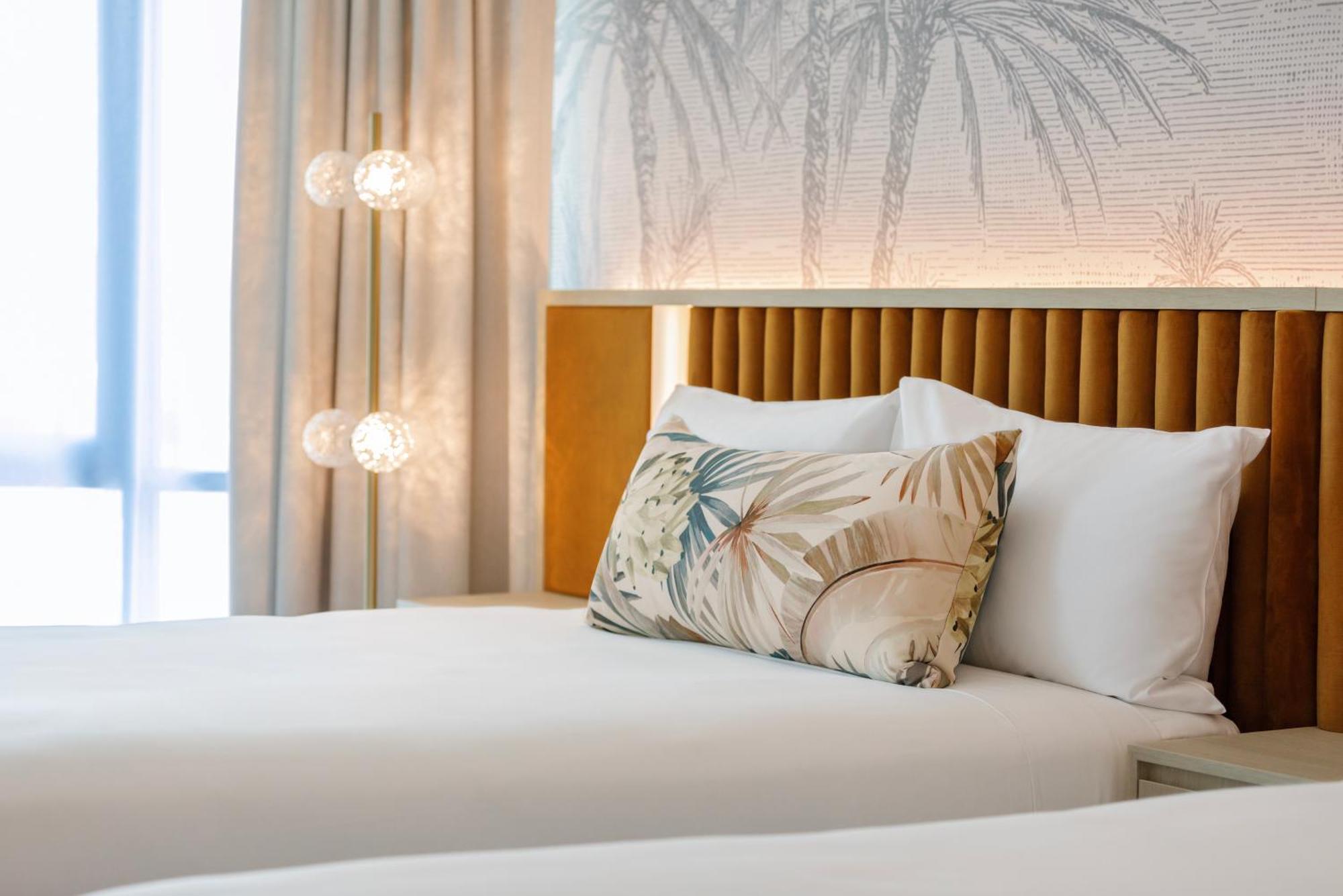 Amora Hotel Брисбен Экстерьер фото The photo shows a stylish hotel room featuring two neatly made beds with white linens. Each bed has several pillows, one of which has a decorative cover with a tropical plant pattern. Behind the beds is a modern headboard, and the wall is decorated w