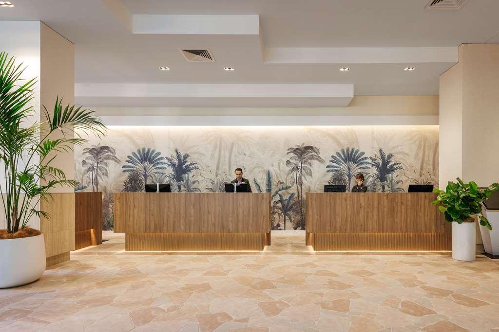 Amora Hotel Брисбен Интерьер фото The photo shows a modern reception area with two wooden reception desks. Behind the desks, there is a decorative wall featuring a large mural of tropical plants and palm trees, creating a serene atmosphere. The space is well-lit with overhead lights,