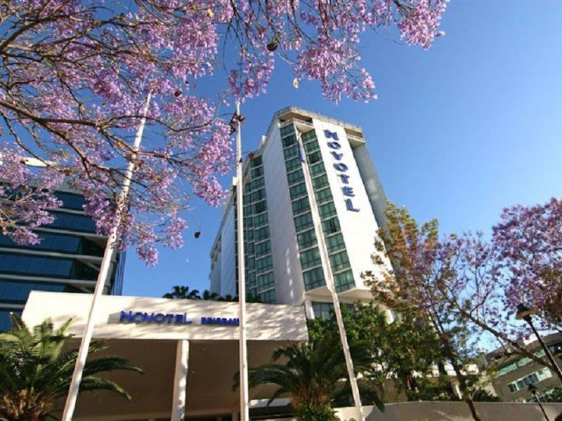 Amora Hotel Брисбен Экстерьер фото The photo shows a modern hotel building, identified as "Novotel," rising above a landscaped area with palm trees. In the foreground, there are vibrant purple flowering trees, likely jacarandas, which add to the picturesque scene. The sky is clear and